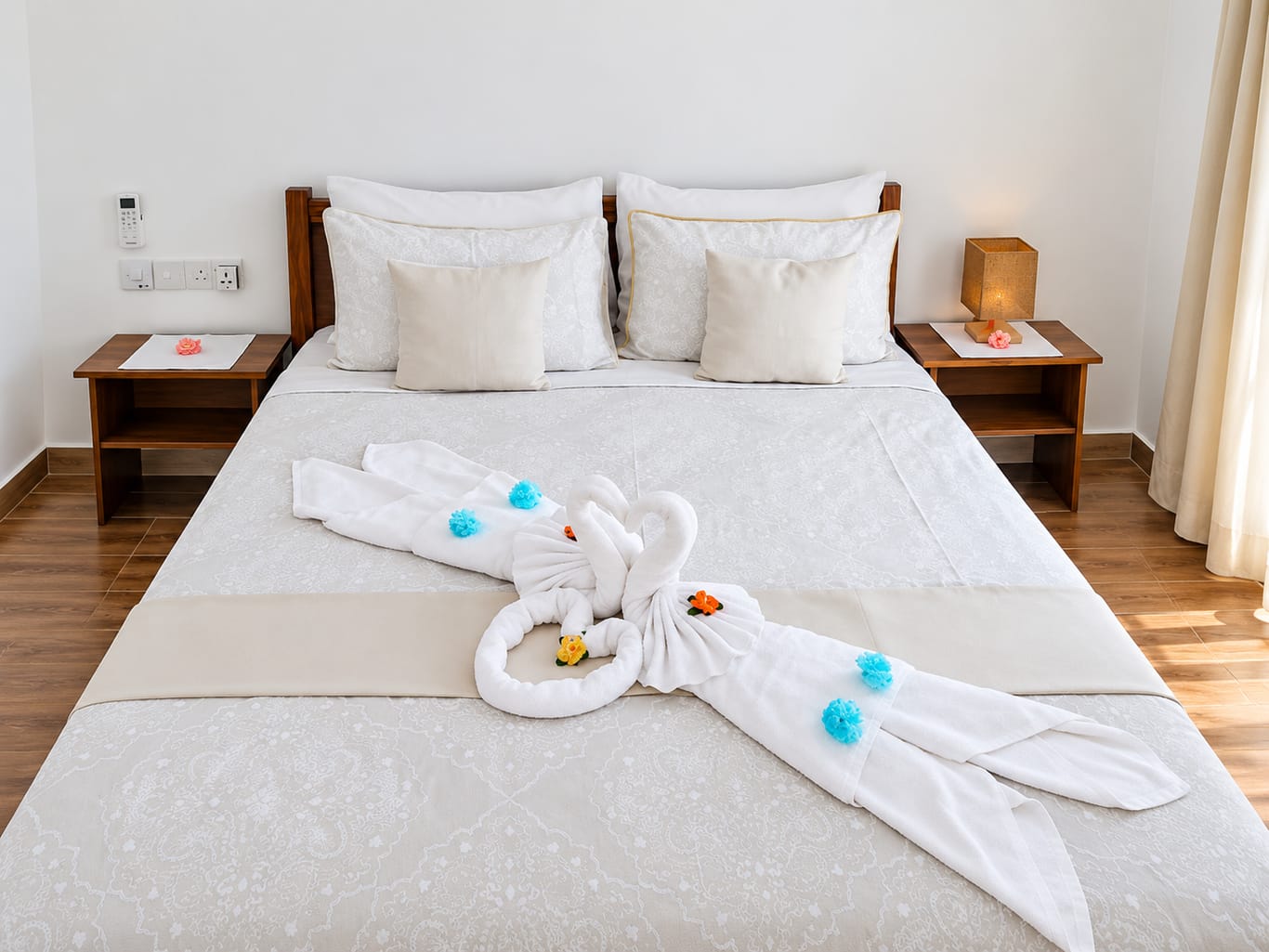 A bedroom in the Two-Bedroom House: a double bed with white patterned linen and towels folded into two swans, with a bedside table on each side.