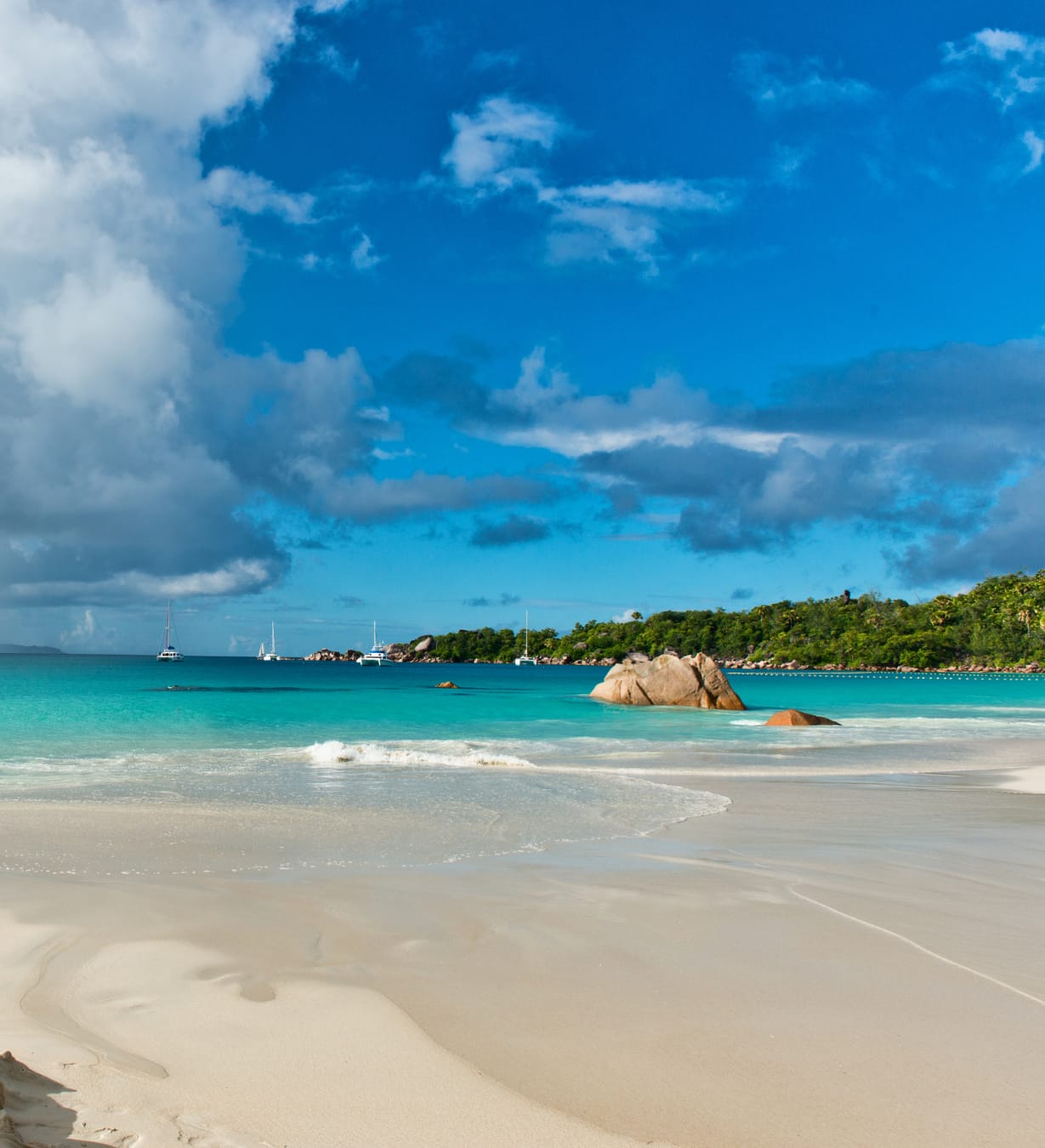 Anse Lazio: pale sand and turquoise water, with granite boulders in the shallows, yachts at anchor and a wooded headland under a blue sky.