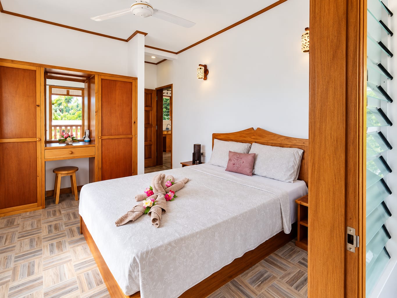 The Apartment bedroom: a double bed with a carved wooden headboard, a ceiling fan, built-in wooden wardrobes and a louvred window.
