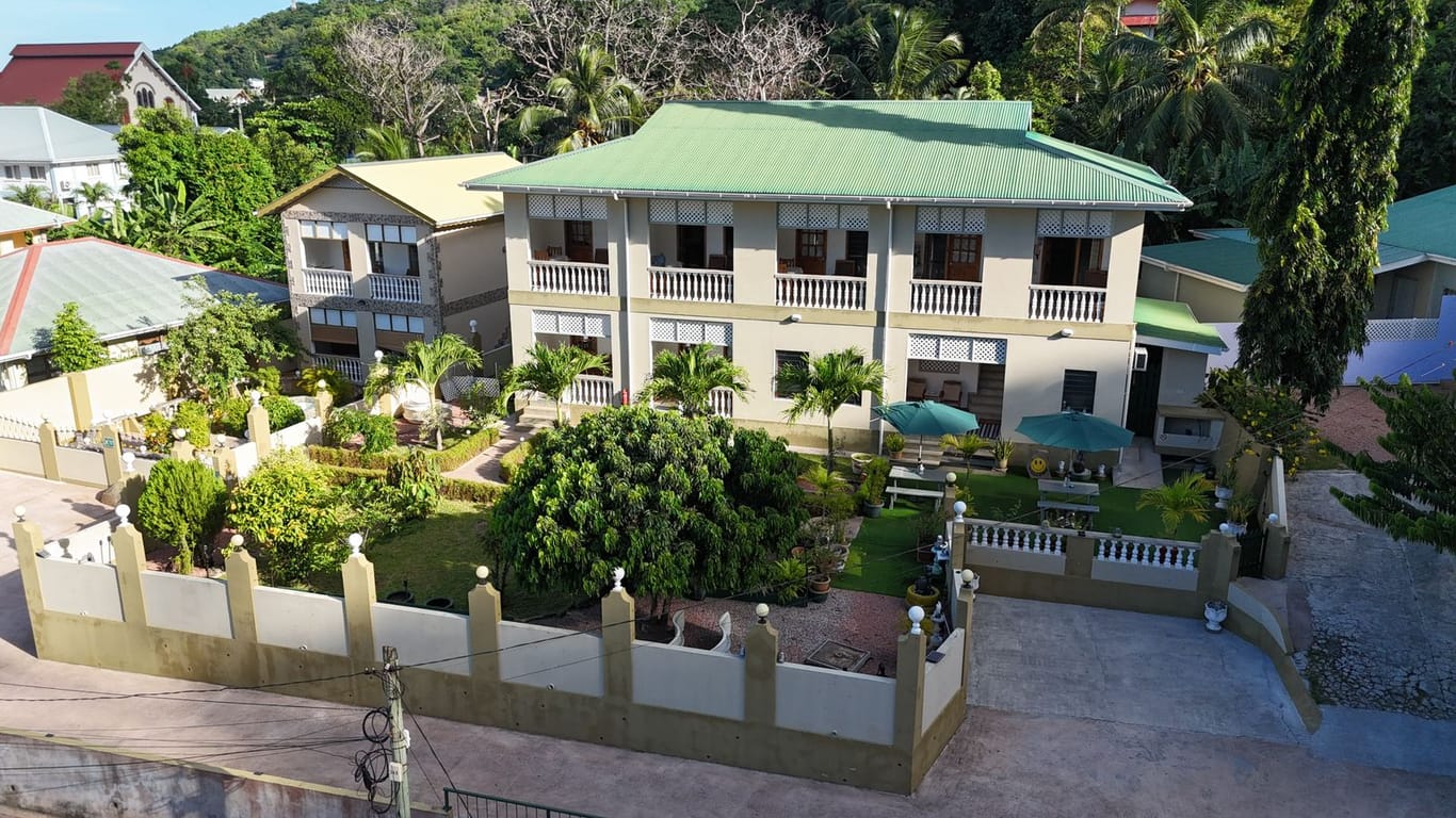 Aerial view of La Residence D'Almée: a two-storey guesthouse with a green roof and white balconies above a palm-planted garden in Baie Sainte Anne, Praslin.