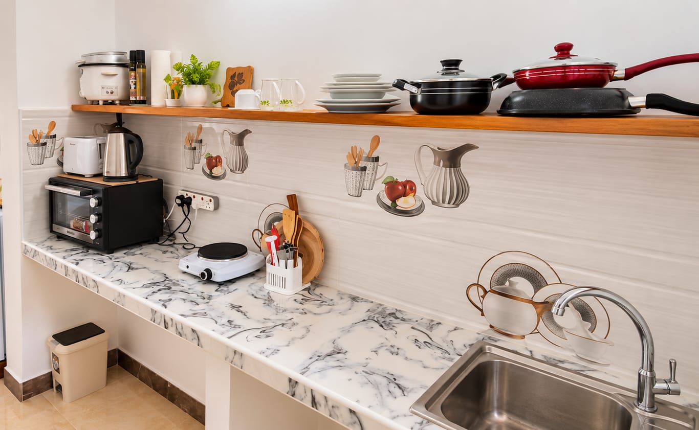 A kitchenette with a marble-effect worktop, sink, electric hotplate, toaster oven, kettle and toaster, with pots, pans and plates on a wooden shelf above.