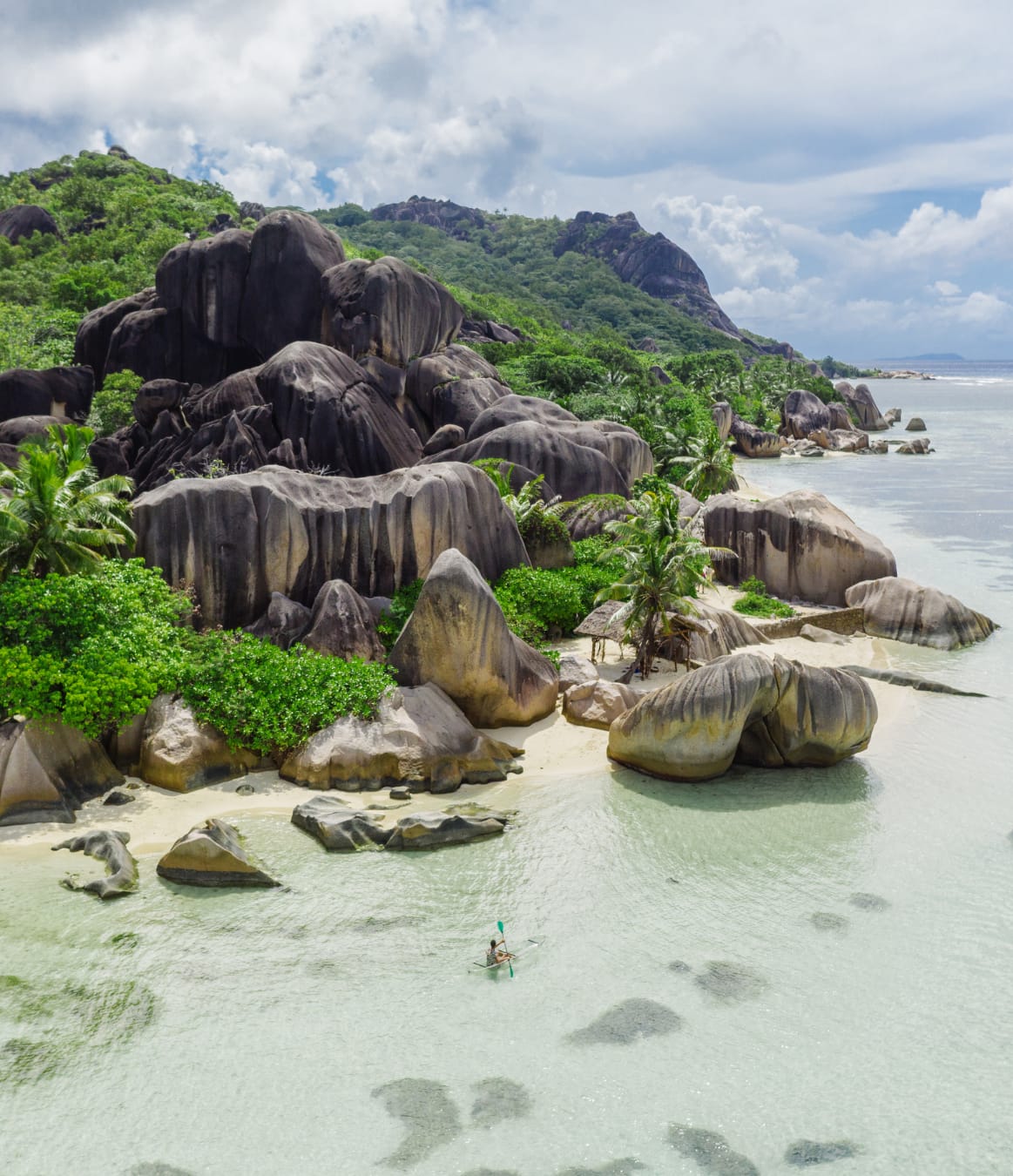Aerial view of a La Digue beach, with giant weathered granite boulders and palms above pale sand and clear shallow water.