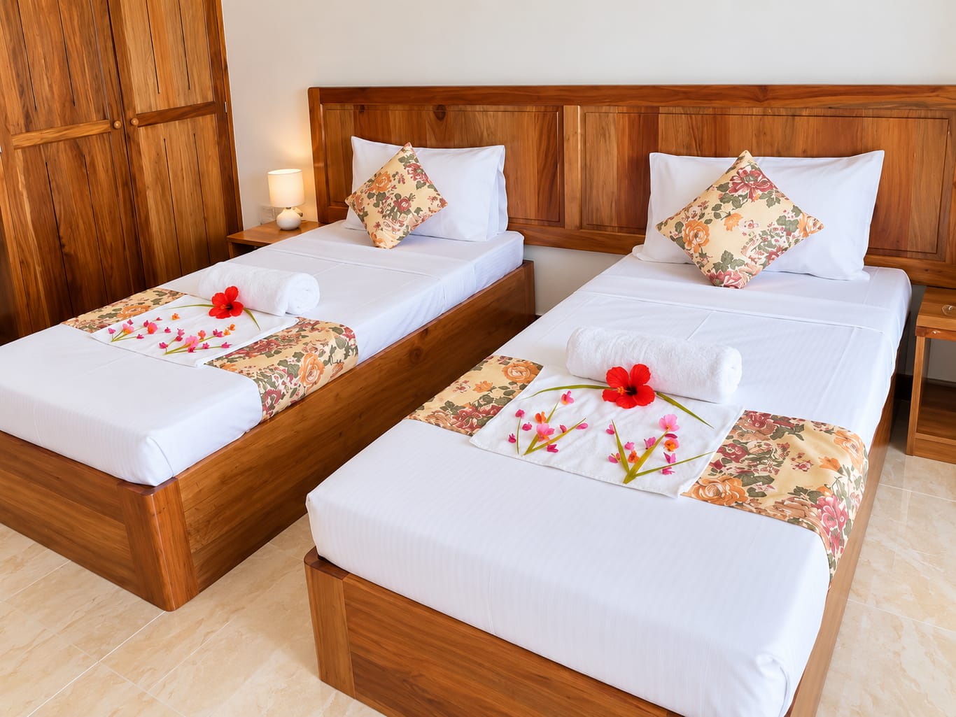A guest room with two single beds in wooden frames, made up with white linen, floral cushions and hibiscus flowers.