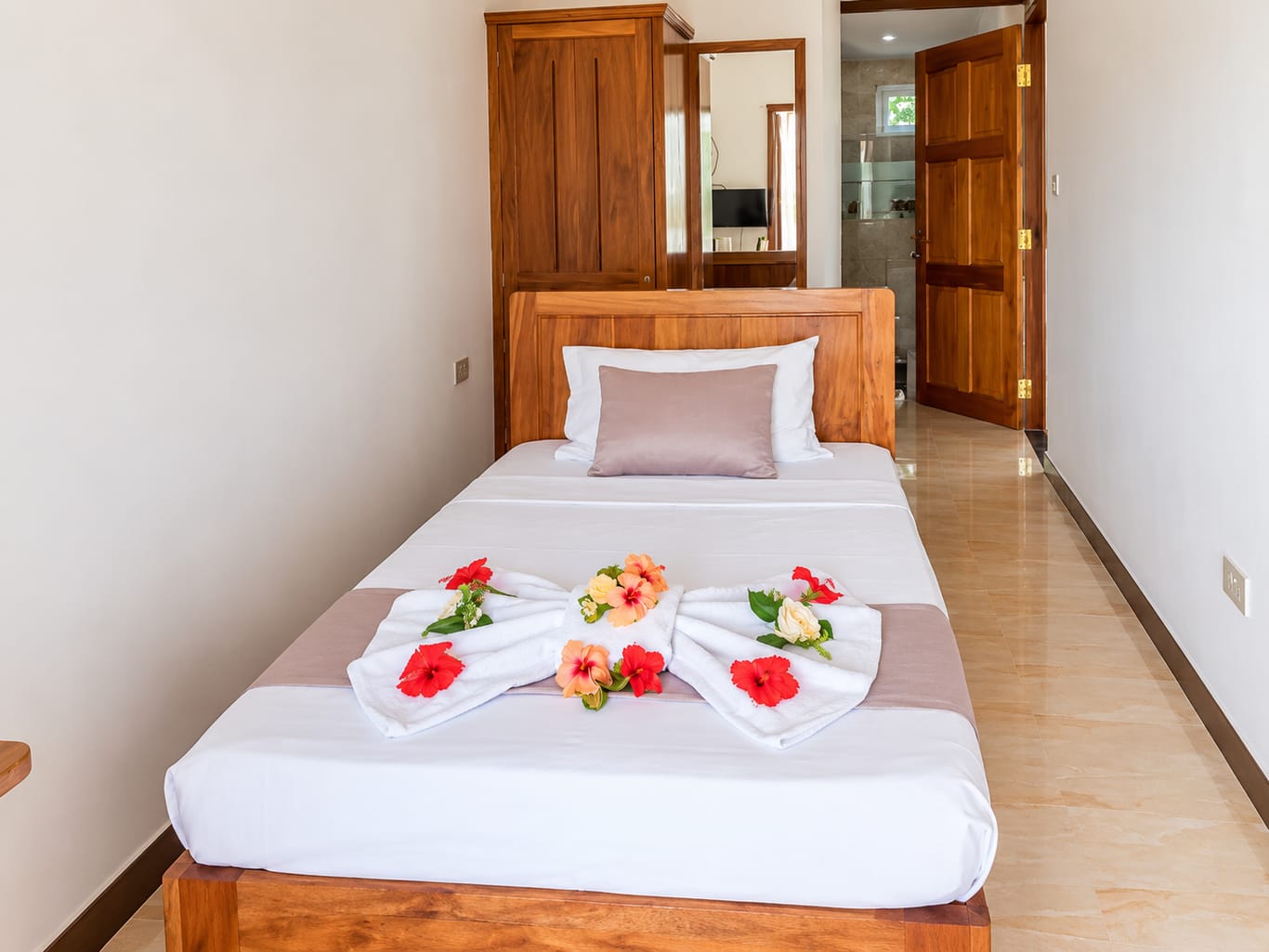 The Single Room: a single bed in a wooden frame with white linen, towels folded into a bow and hibiscus flowers, with a wooden wardrobe and the door to the bathroom behind it.