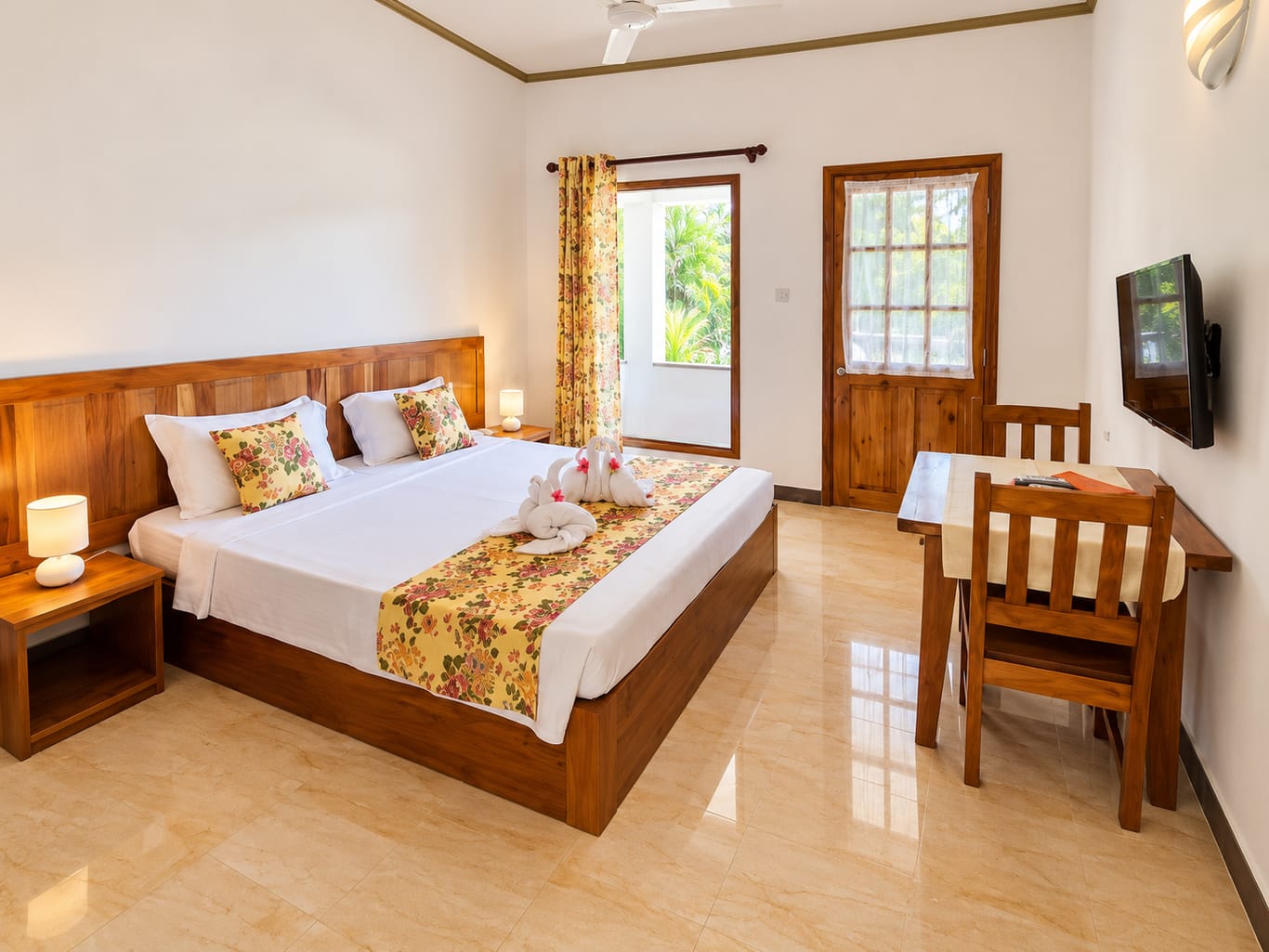 The Standard King Room: a large bed with a wooden headboard, floral cushions and towels folded into two swans, and a table with two chairs beneath a wall-mounted TV.