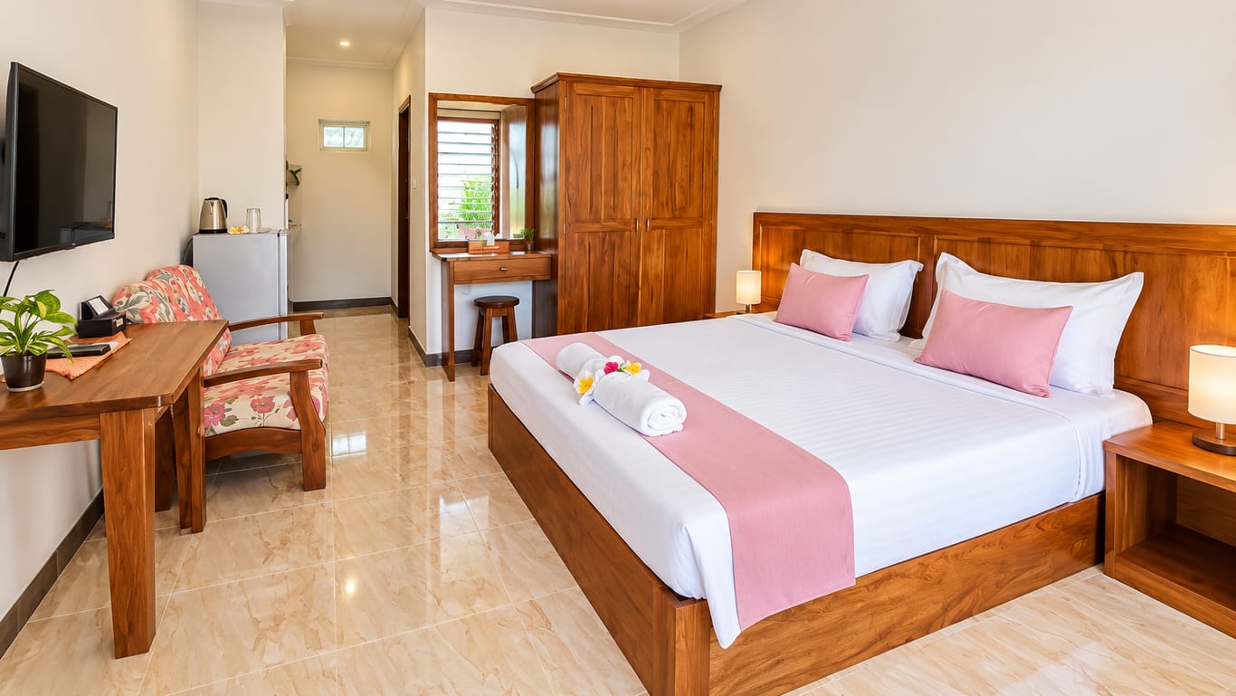 The Studio with Balcony: a large bed with a wooden headboard and pink cushions, a wall-mounted TV, an armchair, a fridge and kettle, and a wooden wardrobe.
