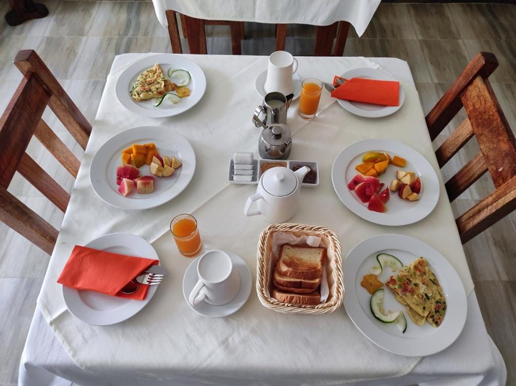Breakfast for two seen from above on a white tablecloth: omelettes, plates of fresh fruit, a basket of toast, orange juice, and pots of tea and coffee.